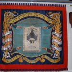 A red and blue banner with Yiddish writing around the outer edge and four hands pointing at the design in the middle