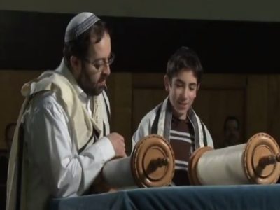 Man with beard in a kippah, glasses and white shirt standing and reading the Torah next to a teenage boy wearing a shawl and also reading the Torah