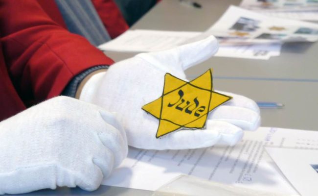 White gloved hand holding yellow star with Jude written on it in Hebrew style letters