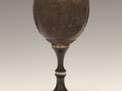 Brown cup made from coconut shell with silver rim. Carved image of men having a meal and using a Kiddush Cup.