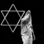 Black and white photo of man covering his face in front of a metal Star of David