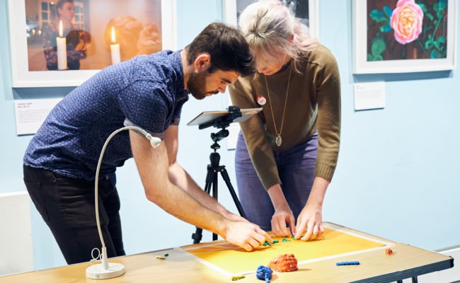 A dark haired man and a blonde woman filming their clay model on a wooden table.