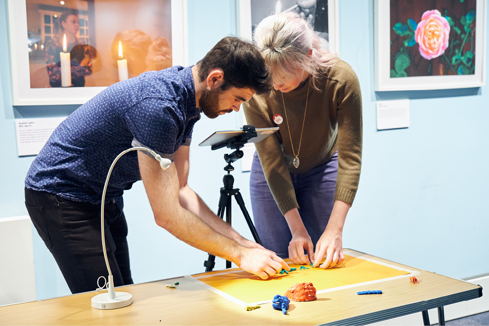 A dark haired man and a blonde woman filming their clay model on a wooden table.