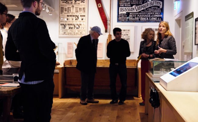Curator giving a tour in the history galleries at the Jewish Museum London in front of a Russian Vapour Baths sign from London's East End