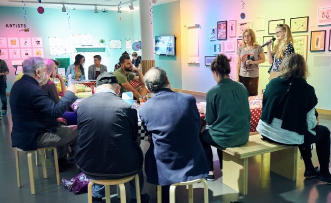 A group of people sitting down and listening to a curator's tour in the Cartoons exhibition at the Jewish Museum London