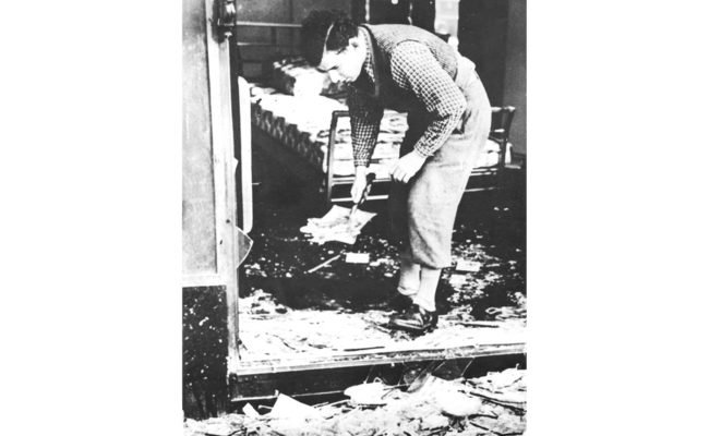 Black and white photograph of adult male bending down to sweep up glass and rubble from vandalised shop doorway