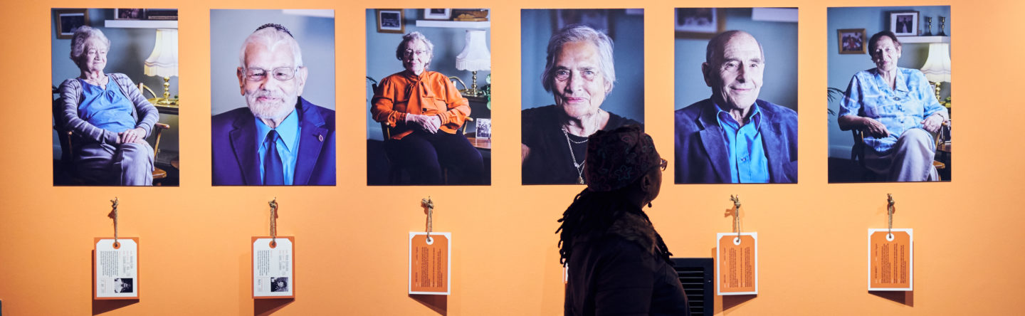 A visitor looks on at 6 colour photographic portaits of the Kindertransport survivors against an orange background