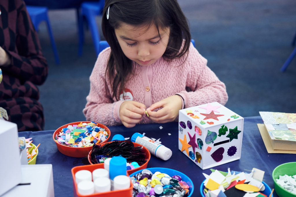 A young girl sitting at a table with blue cloth, glue, and red pots of stickers and gems for a craft activity