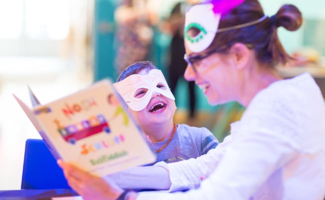 Adult female reads to small child who sis laughing. Both are wearing masks