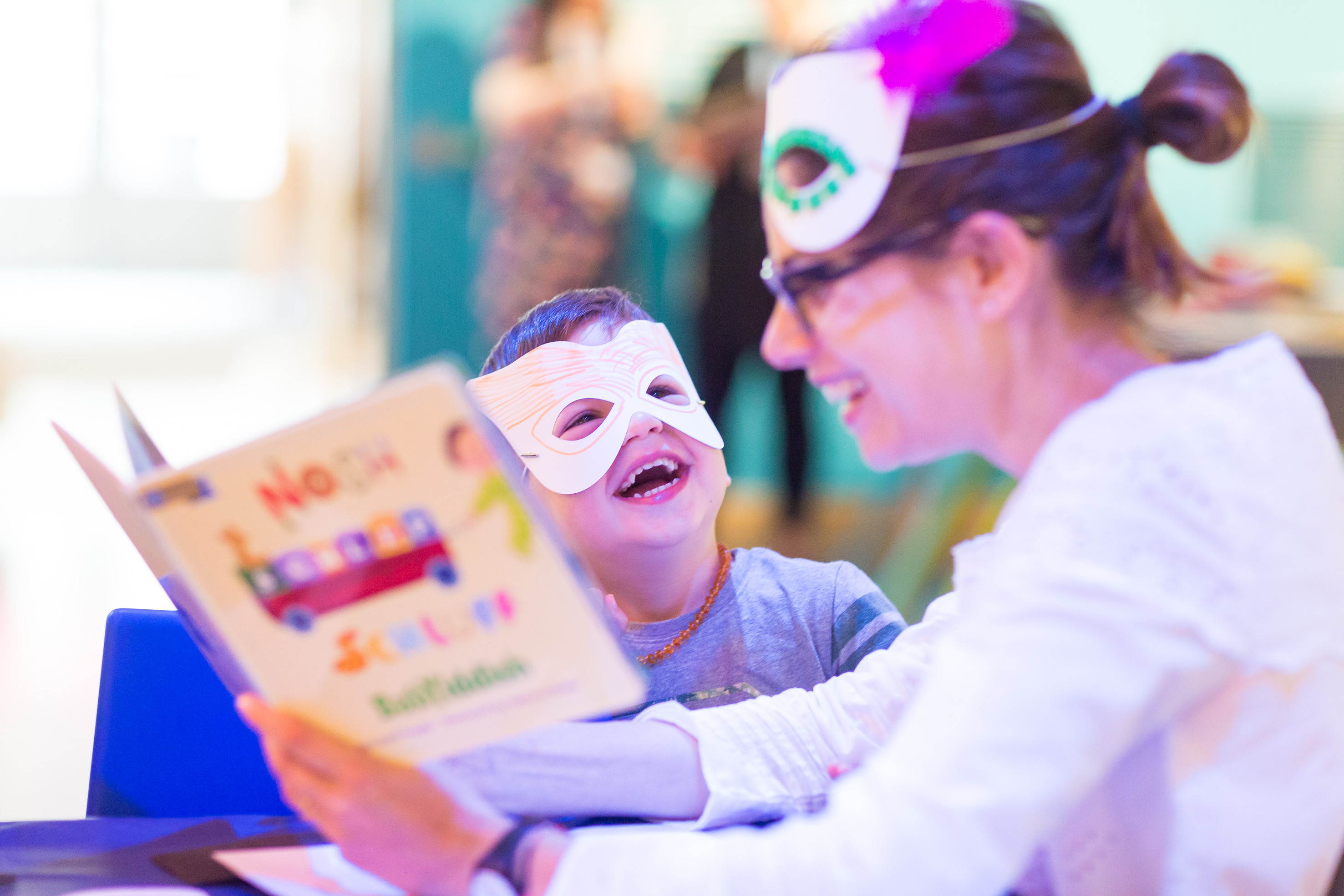 Adult female reads to small child who sis laughing. Both are wearing masks