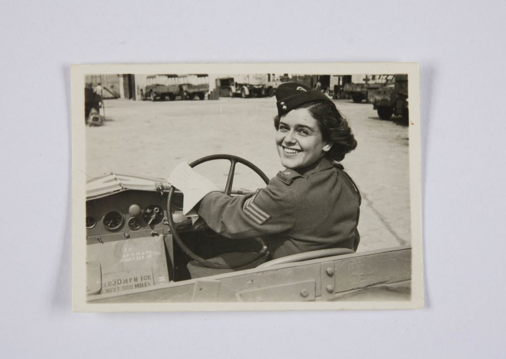 a woman in war time uniform driving an open top car looks back over her shoulder with a smile