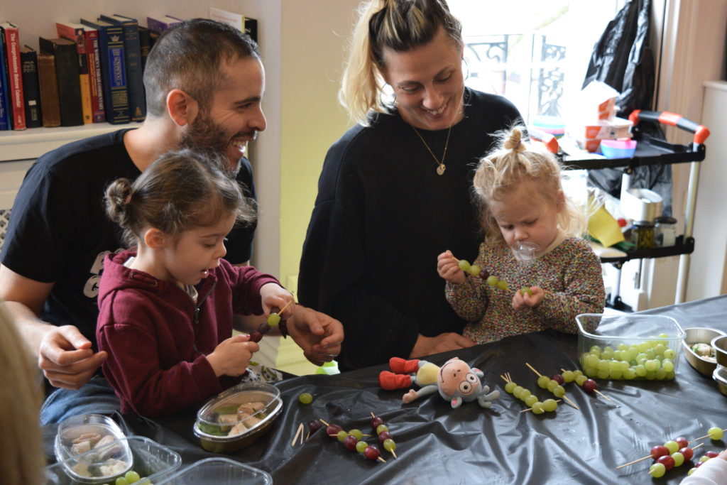 Two adults with children on their laps putting grapes on skewers