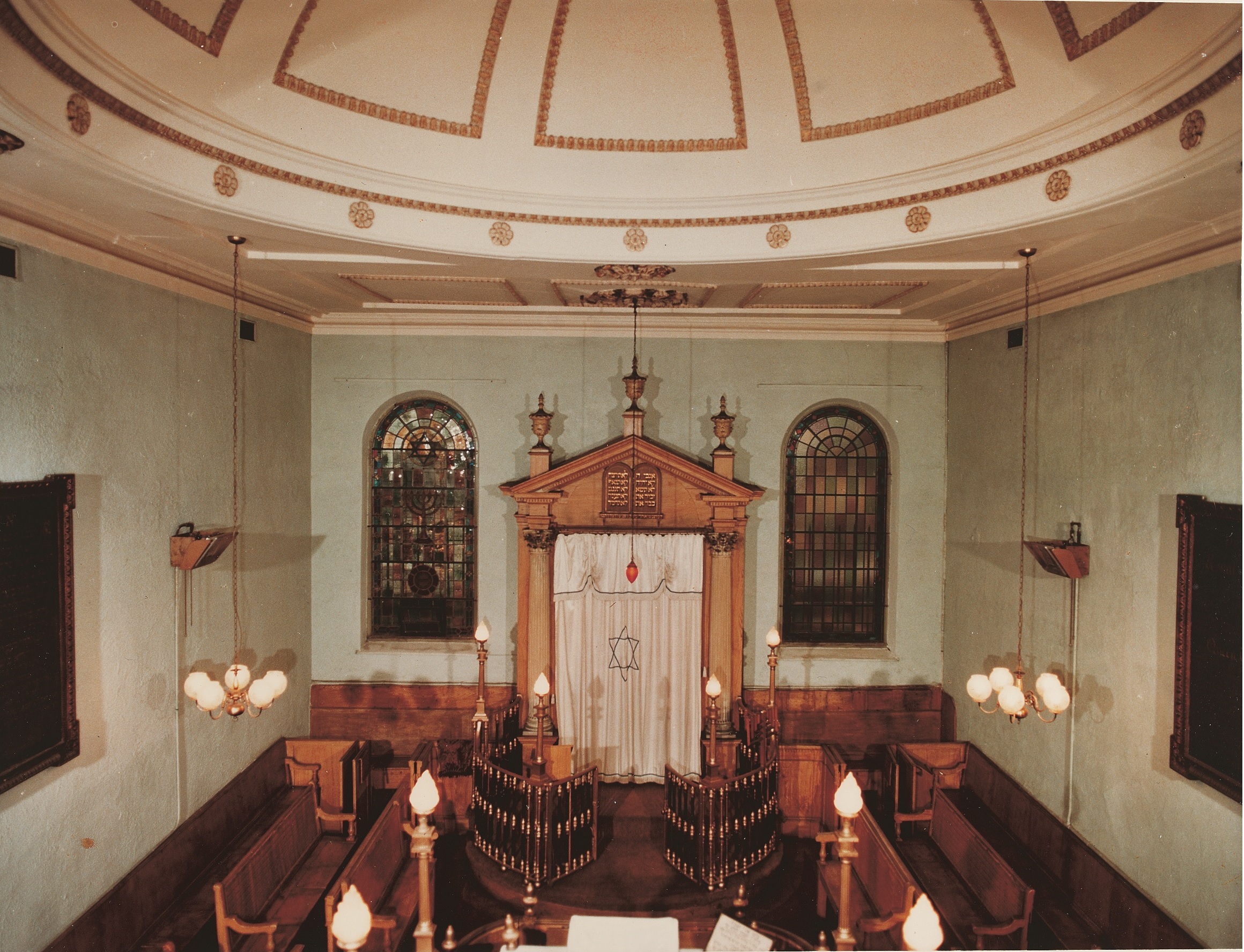 Photograph of synagogue with pale walls, high ceilings, wooden benches and a large white ark.