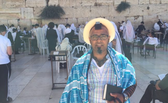 man stands by kotel, wearing blue kente tallit and tefillin