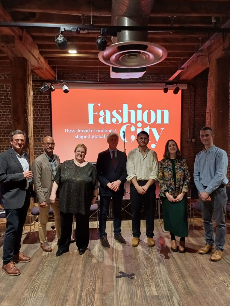 Image of 7 speakers from Jewish Museum London., Museum of the Home, and Museum of London Docklands standing together in front of a backdrop for the Exhibition Fashion City.