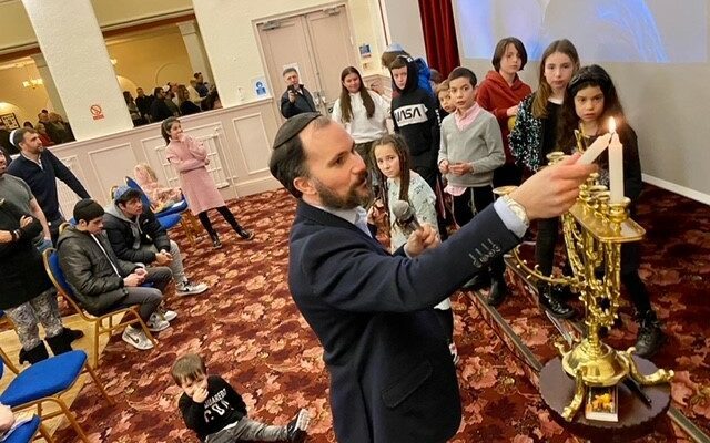shows a Rabbi lighting candles on a Chanukah lamp in front of a group of children